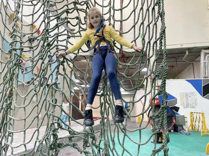 A child, wearing a safety harness, being lowered to the ground with a safety rope in front of a large climbing net, appearing exhausted and looking at the viewer.